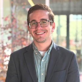 A smiling individual with short brown hair and glasses, dressed in a blue sport jacket, posed against a bright window.