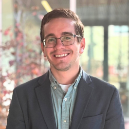 A smiling individual with short brown hair and glasses, dressed in a blue sport jacket, posed against a bright window.
